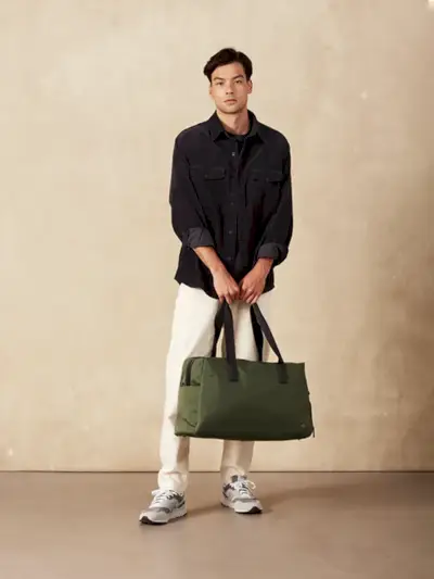 Antler Chelsea Weekender чанта в мек зелен цвят, държана от модел на тъмнокафяв фон