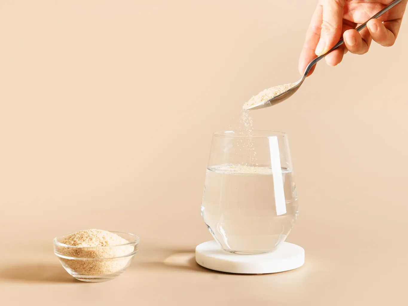 ਇੱਕ ਸੰਗਮਰਮਰ ਕੋਸਟਰ ਉੱਤੇ ਇੱਕ ਚਮਚੇ ਤੋਂ ਇੱਕ ਗਲਾਸ ਪਾਣੀ ਵਿੱਚ ਫਾਈਬਰ ਪੂਰਕ ਪਾਊਡਰ ਛਿੜਕਦੇ ਹੋਏ ਹੱਥ
