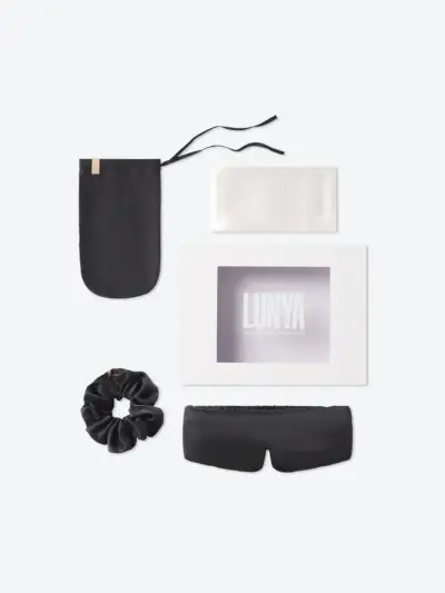 Lunya Sleep The Details mit verschiedenen Schlafaccessoires aus schwarzer Seide neben einer Markenverpackung auf grauem Hintergrund